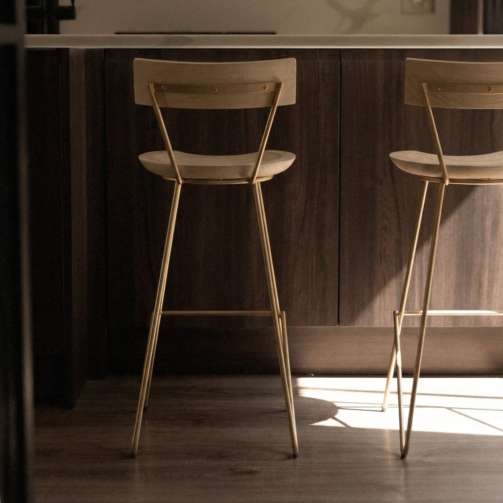 Two modern bar stools with light beige upholstered seats and backs, supported by slim gold metal legs, positioned in front of a dark wood kitchen island.