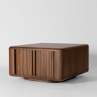 A mid-century modern wooden coffee table with a warm brown finish, featuring vertical slatted front doors and a rounded top, photographed on a light grey studio background.